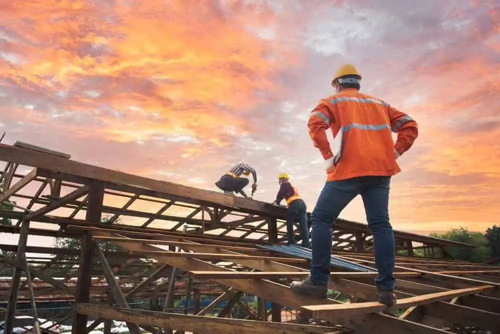 Construction workers wearing safety gear work on a wooden structure at sunset. One stands, overseeing the progress, while two others crouch and work on the beams. The sky is filled with vibrant orange and pink hues.