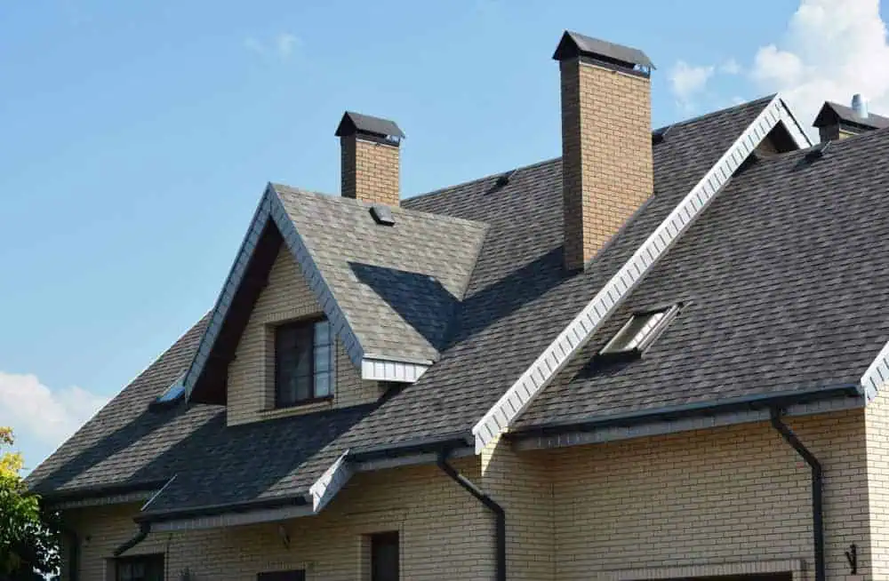 A modern house with a sloped roof covered in gray shingles. The house features two brick chimneys, triangular dormer windows, and beige brick walls. The sky is blue with a few clouds.