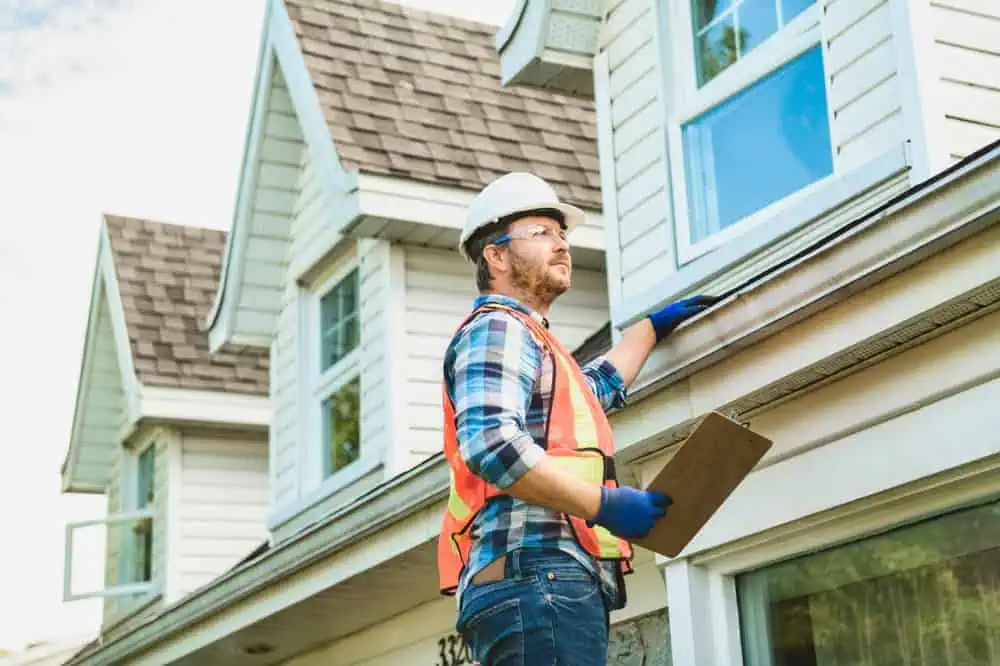 A construction worker wearing a hard hat, safety vest, and gloves is inspecting the gutter of a house. He holds a clipboard and stands on a ladder beside the house, which has white siding and a shingled roof.