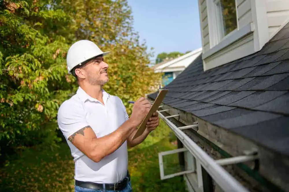 A man wearing a white hard hat and polo shirt is inspecting a house's gutter while holding a clipboard. He stands next to a shingled roof, with green trees in the background under a clear sky.