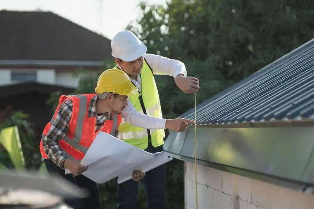 Two construction workers wearing hard hats and safety vests are inspecting the edge of a roof. One is holding a blueprint, while the other is measuring with a tape. They are outdoors, and trees are visible in the background.