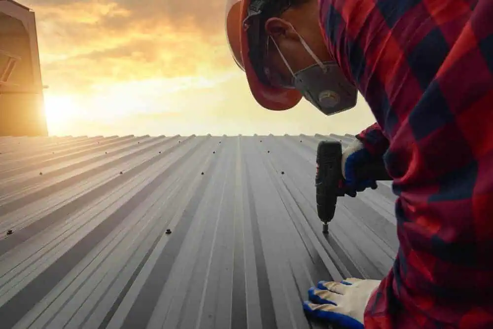 A construction worker wearing a red checkered shirt, helmet, mask, and gloves uses a power drill on a sheet metal roof as the sun sets in the background.