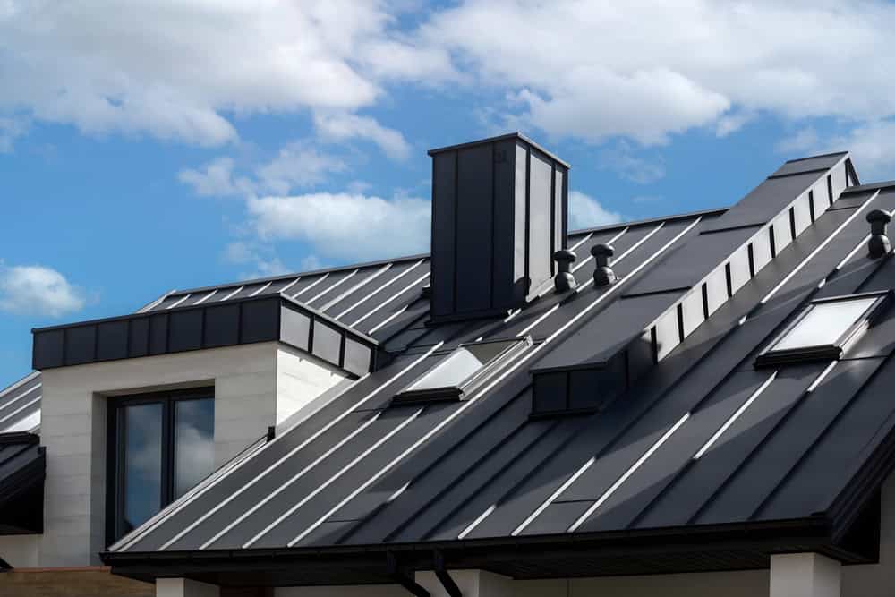 A modern building with a sleek black metal roof featuring skylights and a chimney against a blue sky with scattered clouds.