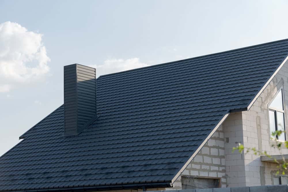A house with an unfinished, white brick exterior features a newly installed dark shingled roof and a modern chimney against a clear blue sky.