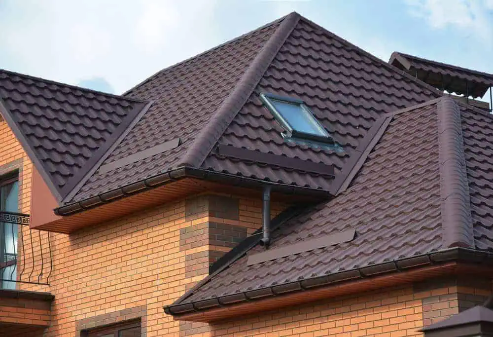 A brick house features a complex, dark brown metal roof with multiple gables. A skylight is visible, and the roof's textured tiles highlight its modern design. The bricks alternate between brown and red shades, adding to the architectural detail.
